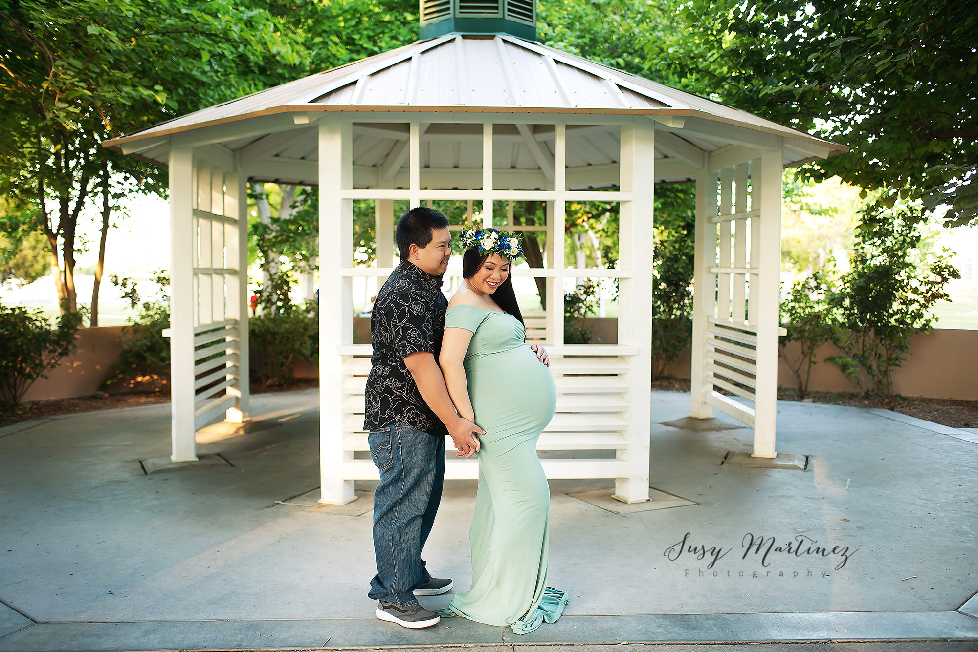 mom in light green maternity gown with flower crown poses by gazebo with husband