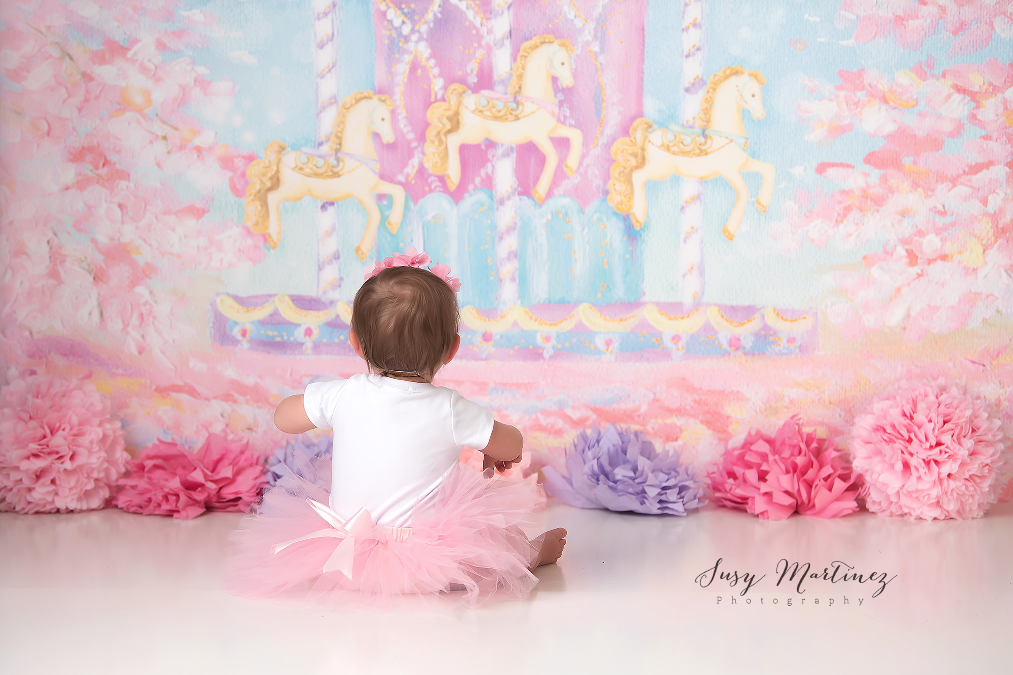 baby girl looks at carousel on backdrop during milestone portraits for first birthday