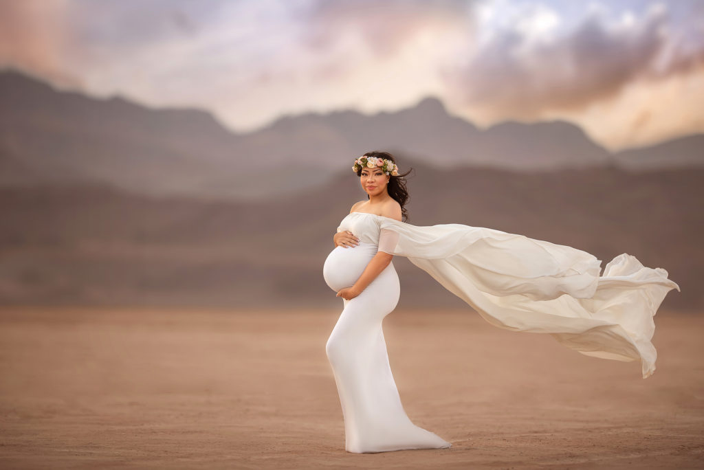 mother in ivory gown holds stomach while wind blows wrap during Dry Lake Bed maternity session