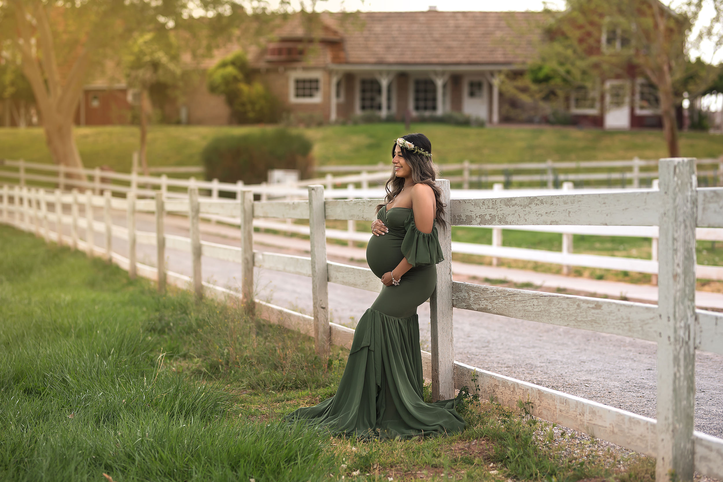 woman in green off-the-shoulder gown leans against white fence during Spring Mountain Ranch Maternity Session