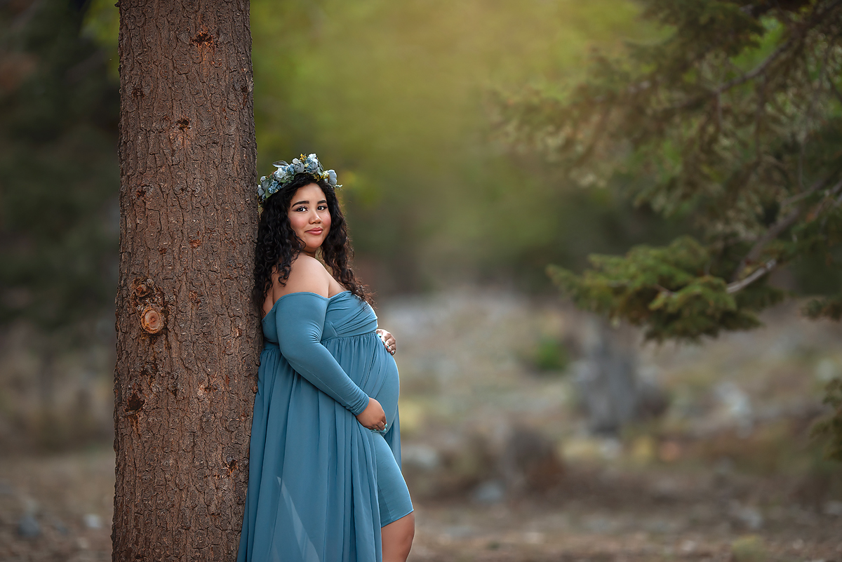 expecting mother leans against tree in blue Sew Trendy accessories gown during maternity portraits 